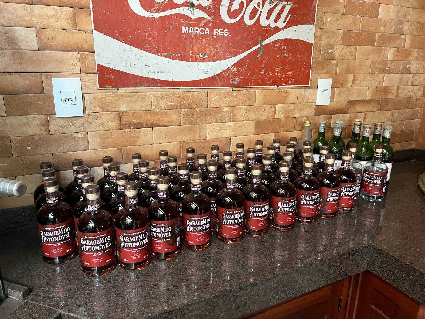 Garrafas da Cachaça Garagem do Automóvel sobre bancada de granito, com placa Coca-Cola vintage ao fundo
